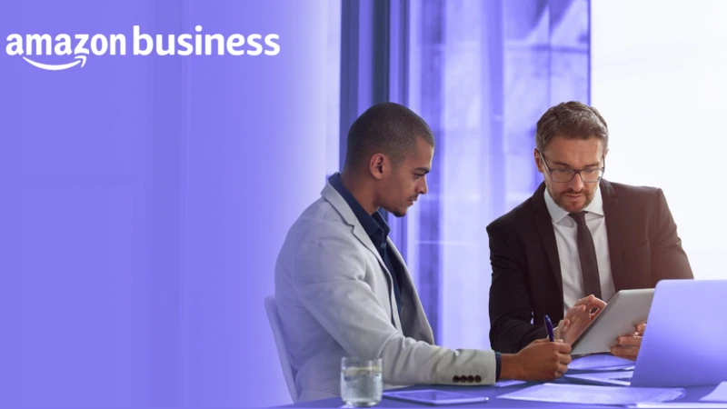 Two business professionals engaged in a discussion at a modern office, with one using a tablet and the other taking notes. The image features the "Amazon Business" logo in the top left corner, symbolizing strategic business procurement and collaboration.